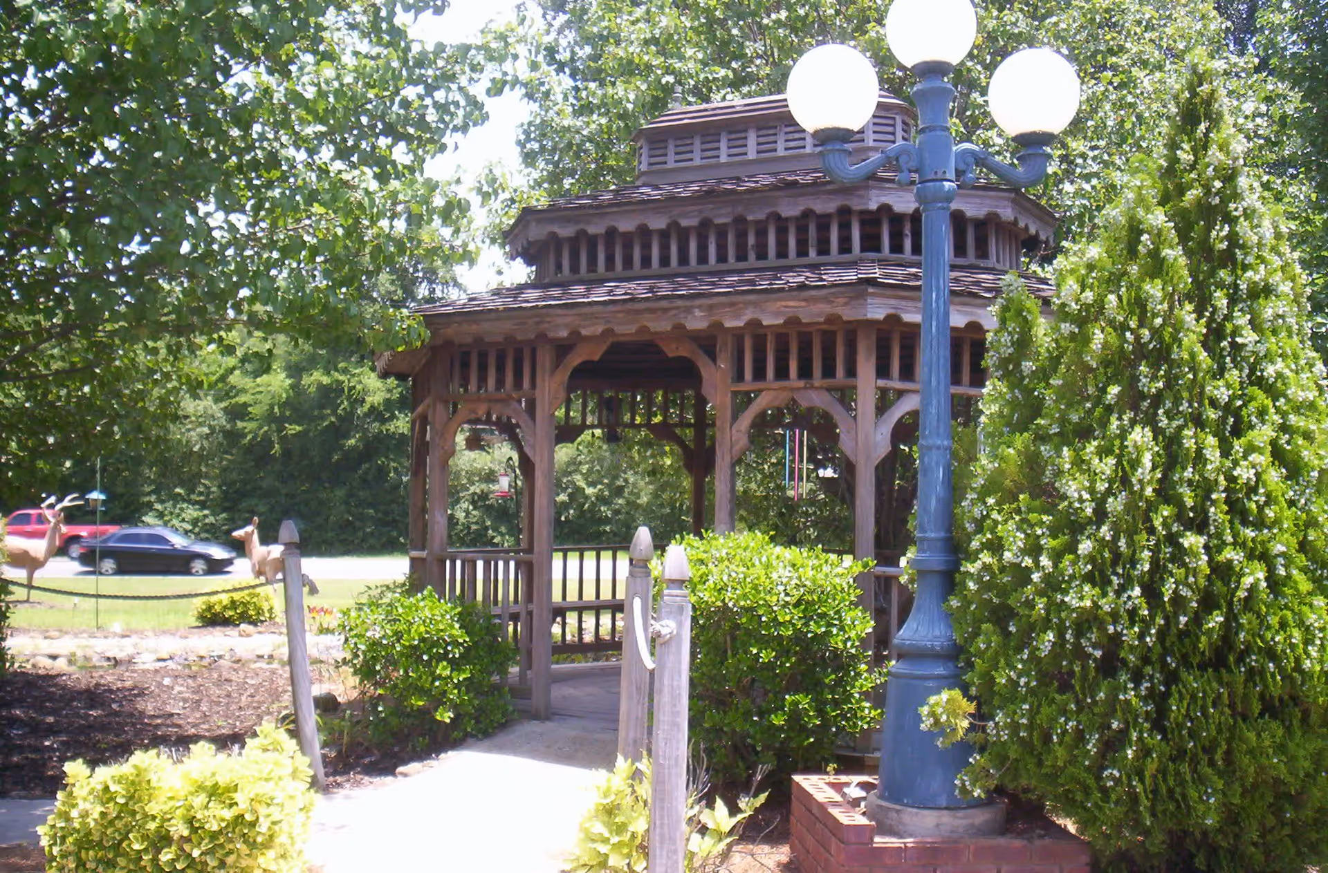 A wooden gazebo surrounded by green bushes and trees in a garden area with a blue street lamp featuring three round white lights nearby. In the background, there are two cars on a road and some deer statues on the grass.
