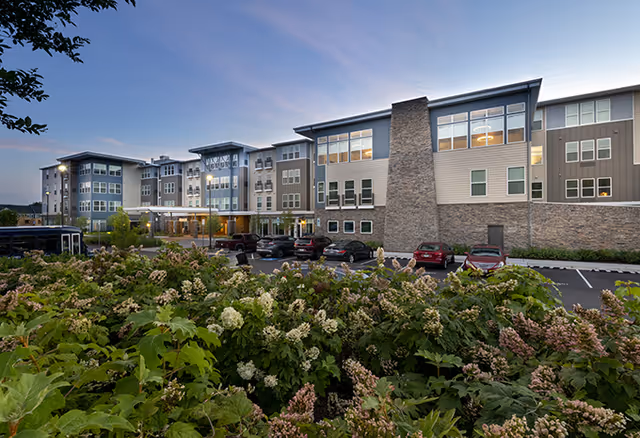 Exterior front view of a multi-story senior living building at dusk with landscaped shrubs and a parking lot.