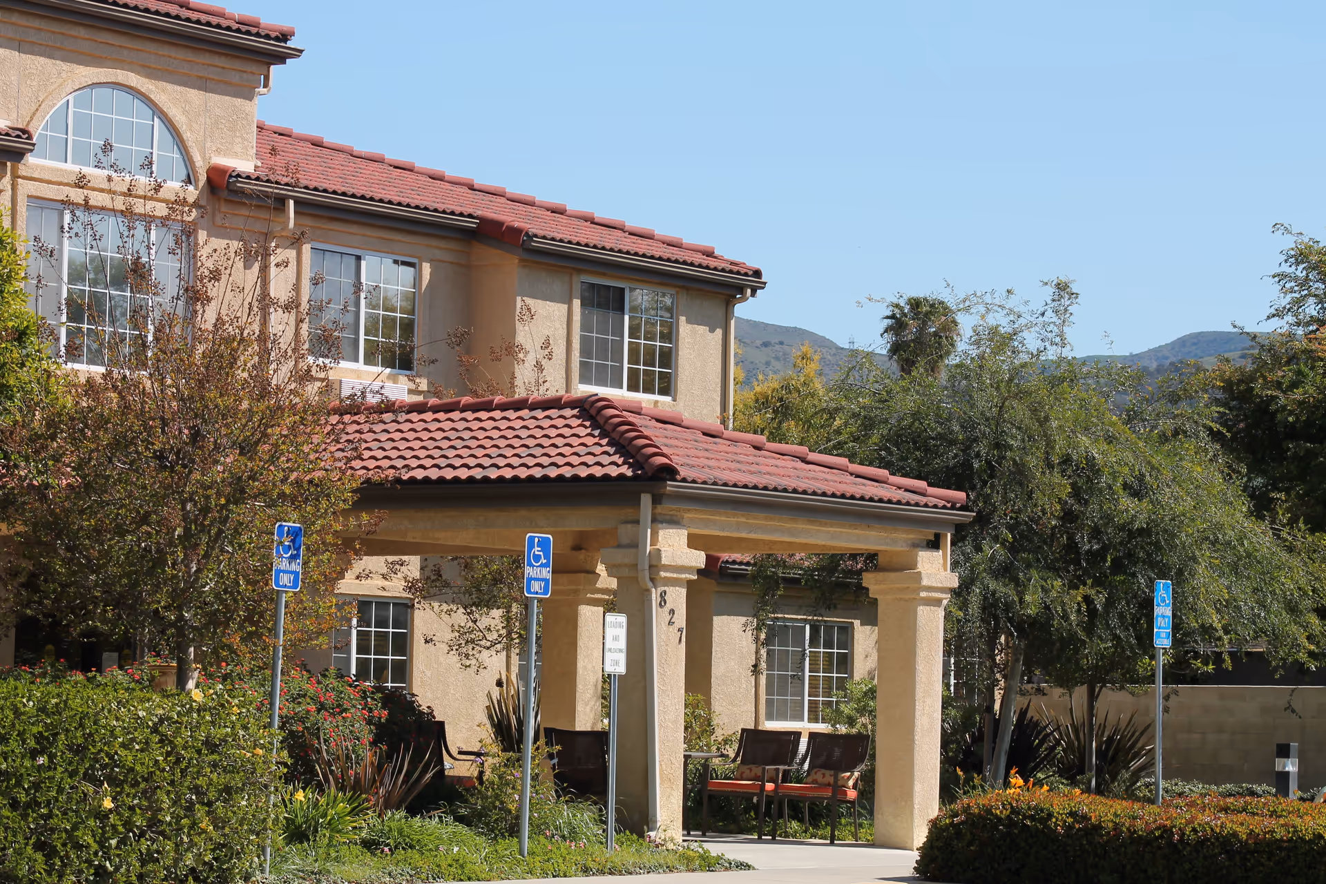 Exterior view of a senior living facility building with beige stucco walls and red tiled roof. The entrance has a covered porch supported by columns, with benches underneath. There are several handicap parking signs visible near the entrance. Surrounding the building are trees, bushes, and landscaped greenery with mountains in the background under a clear blue sky.