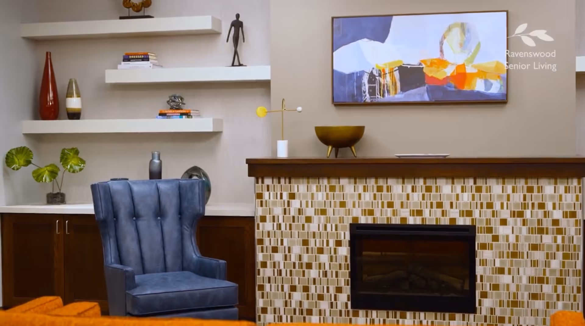 A cozy living room area featuring a blue upholstered armchair, a fireplace with a mosaic tile surround, and a wooden mantel. Above the mantel is a colorful abstract painting. To the left, there are three white floating shelves with decorative items including books, vases, and a small sculpture. The room has a warm and inviting atmosphere.