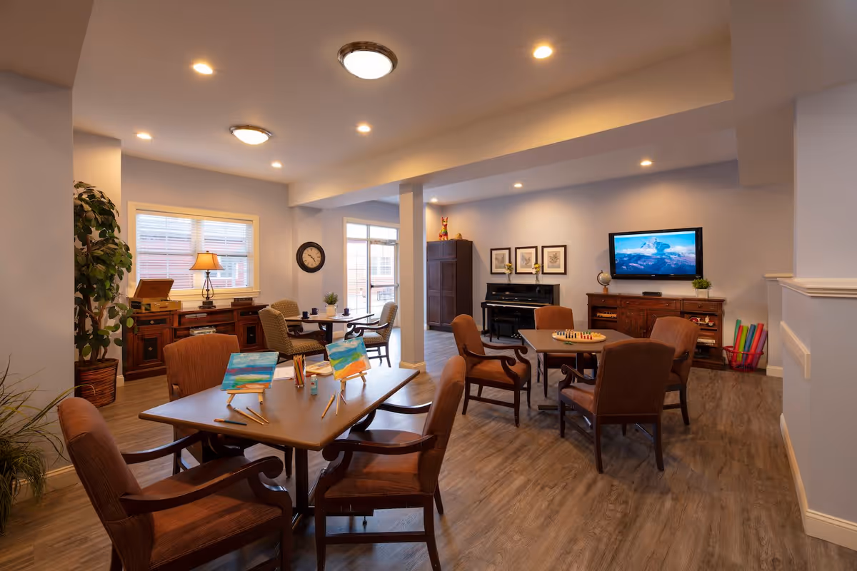 A well-lit activity room in a senior living facility with several tables and chairs arranged for group activities. One table has small easels with colorful paintings and art supplies, while another table has a board game set up. The room features wooden furniture, a piano against the wall, a wall-mounted TV, framed artwork, and a clock. There are large windows and a door letting in natural light, and the floor is covered with wood-style flooring.
