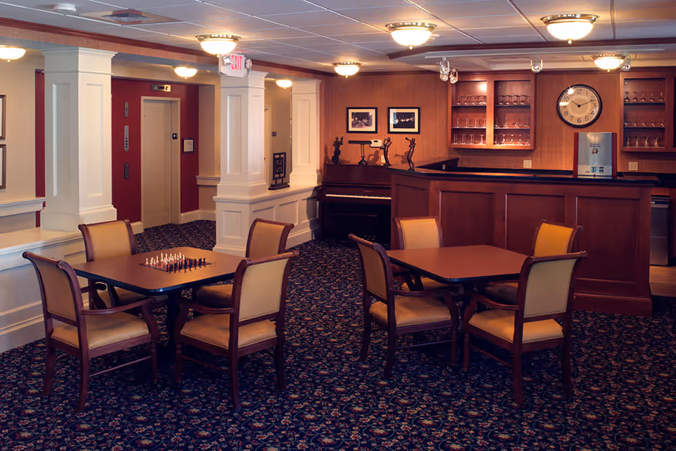 A cozy common area in a senior living facility featuring two tables with chairs, one table set with a chessboard. The room has patterned carpet, white pillars, a piano against the wall, and a wooden bar with glass shelves and a clock above it. Warm lighting fixtures illuminate the space.