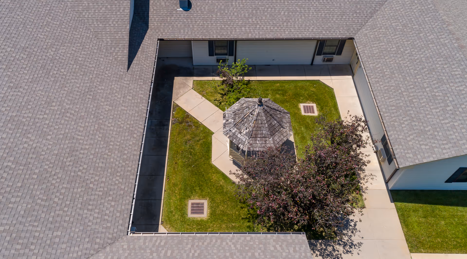 Aerial view of a courtyard in a senior living facility showing a small wooden gazebo surrounded by green grass, a tree with purple leaves, and concrete walkways. The courtyard is enclosed by the building's roof and walls.