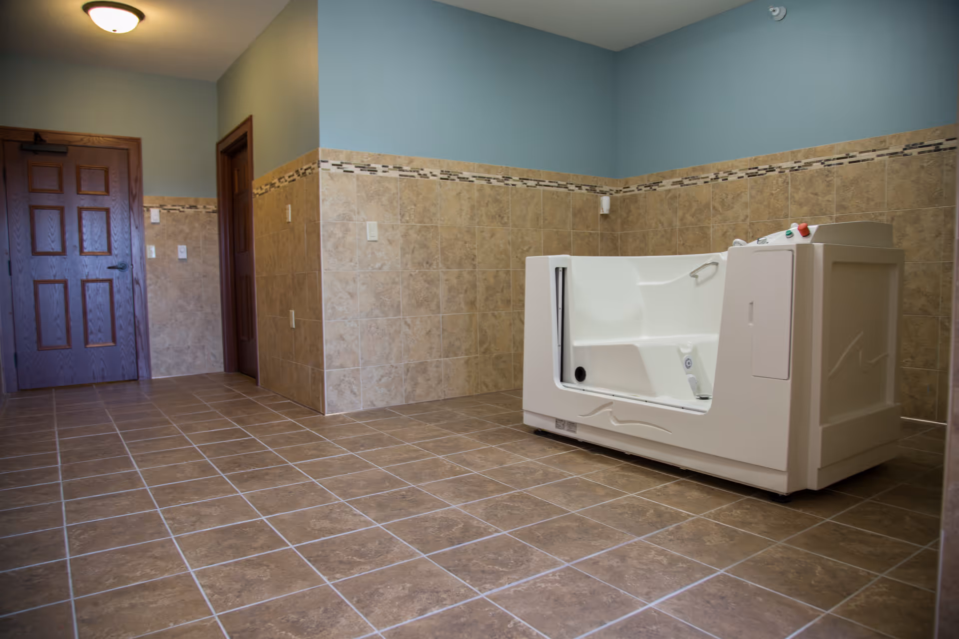 A spacious room with tiled floor and walls partially covered with beige tiles and a decorative border. The room contains a white walk-in bathtub with control buttons on the top edge. There are two wooden doors on the left side of the room and a ceiling light fixture.