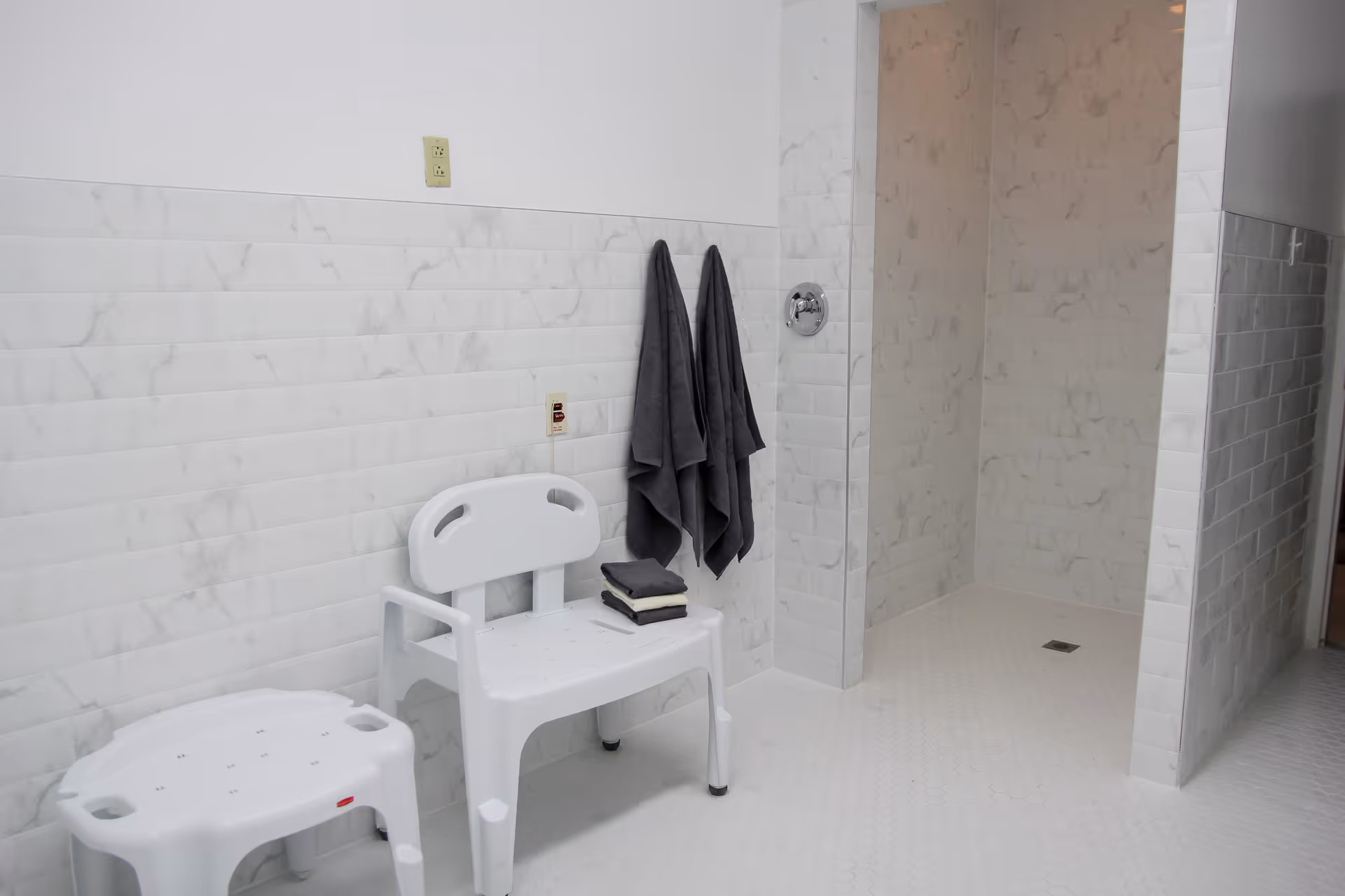 Tiled accessible shower room with a walk-in shower, white plastic shower bench and stool, and towels hanging on the wall.