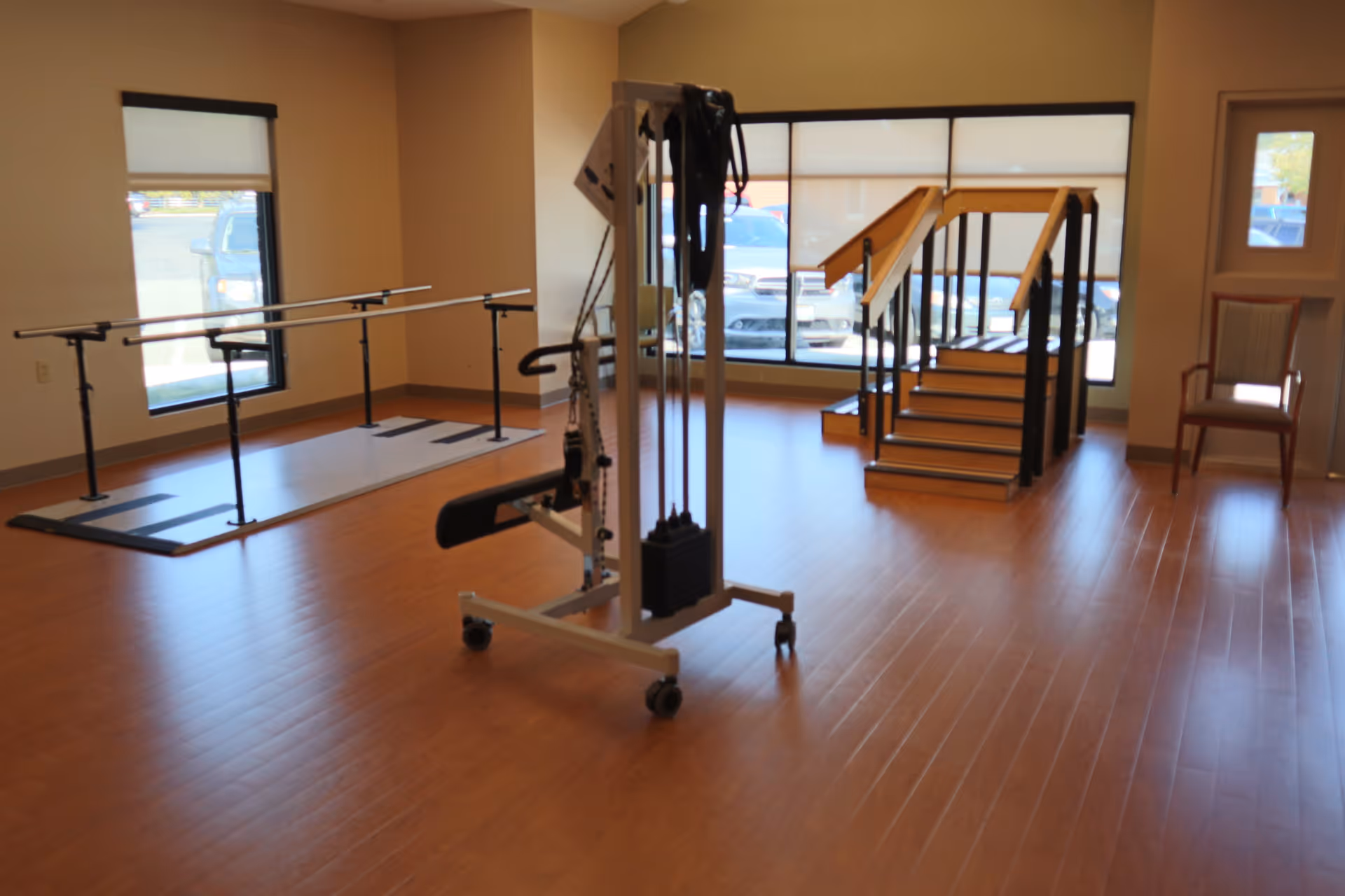 A rehabilitation or physical therapy room with wooden flooring, parallel bars for walking exercises, a set of stairs with handrails for stair climbing practice, and a piece of exercise equipment in the center. Large windows allow natural light to enter the room.