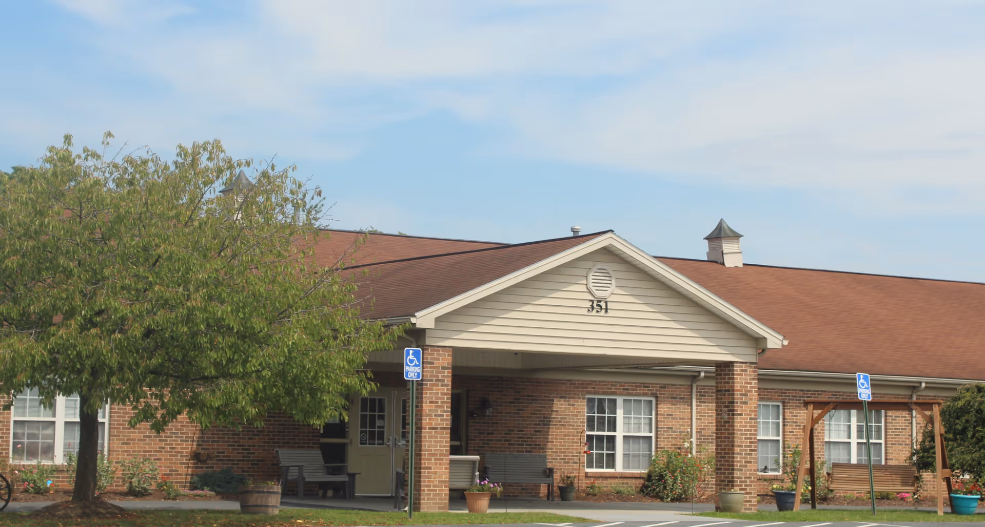 Exterior view of a single-story brick building with a brown roof and a covered entrance. There are benches near the entrance, a tree on the left side, and two handicap parking signs visible in front of the building. The building number 351 is displayed on the front gable.