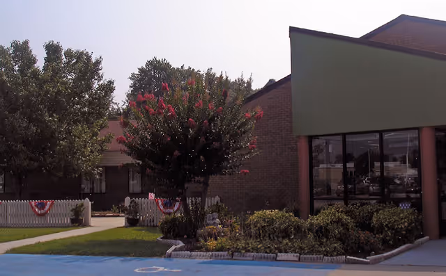 Front exterior of a nursing facility showing a green-awned entrance with large windows, a flowering tree, white picket fence with patriotic bunting, and landscaped parking area.