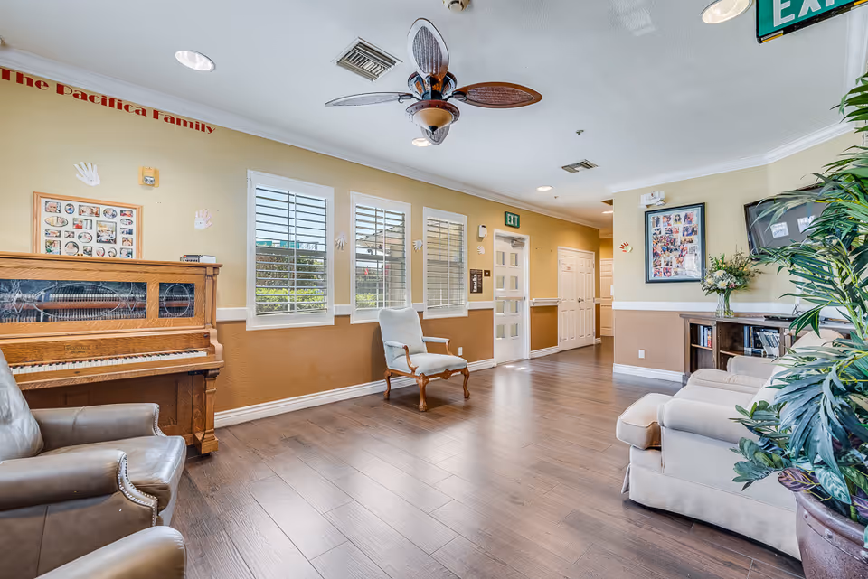 Bright common lounge with chairs, a piano, plants, and windows in a senior living facility.