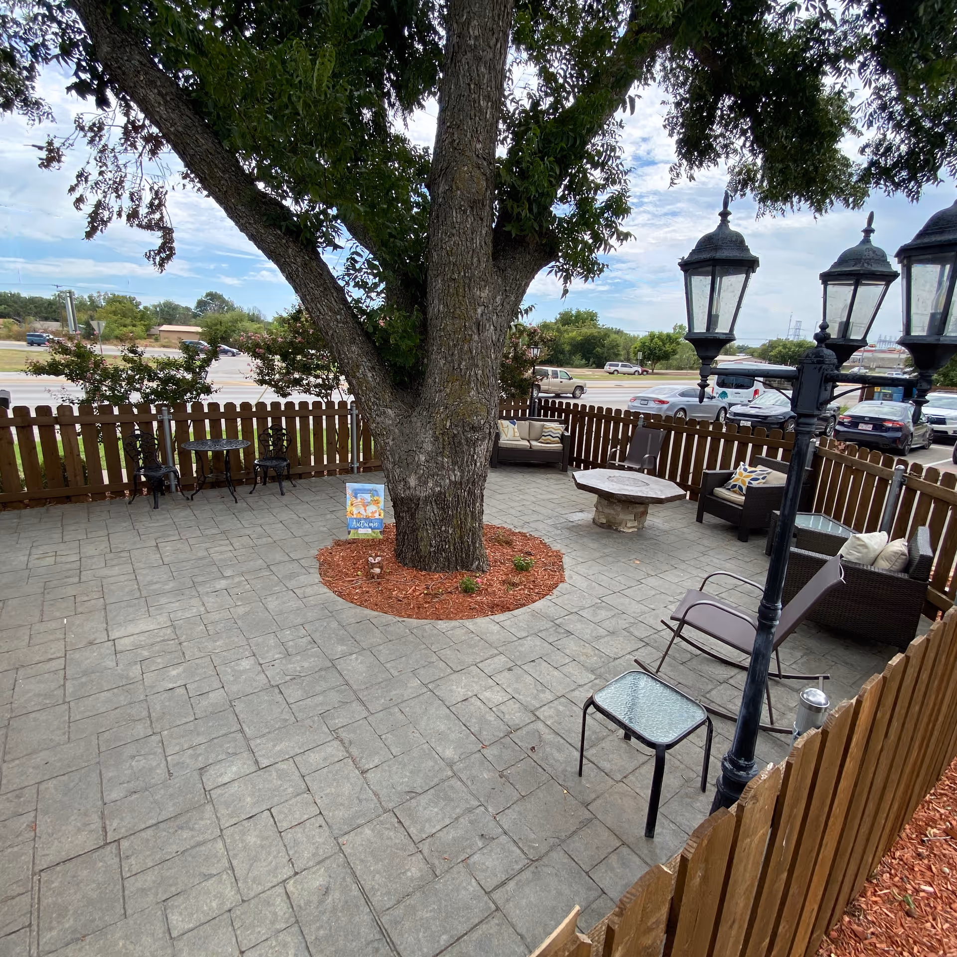 Outdoor patio area with a large tree in the center surrounded by a circular mulch bed. The patio is paved with stone tiles and enclosed by a wooden fence. There are several seating arrangements including metal chairs with a small round table, cushioned wicker chairs, and a stone fire pit. A vintage-style black street lamp is also visible. In the background, cars are parked along a road with trees and buildings further away under a partly cloudy sky.