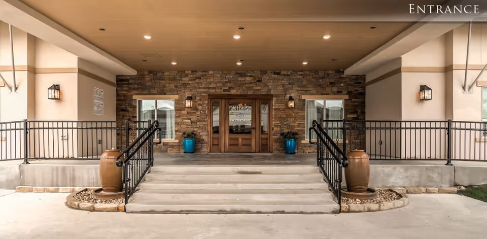 Covered entrance with stone facade and double wooden doors accessed by steps with black railings and large planters on either side.