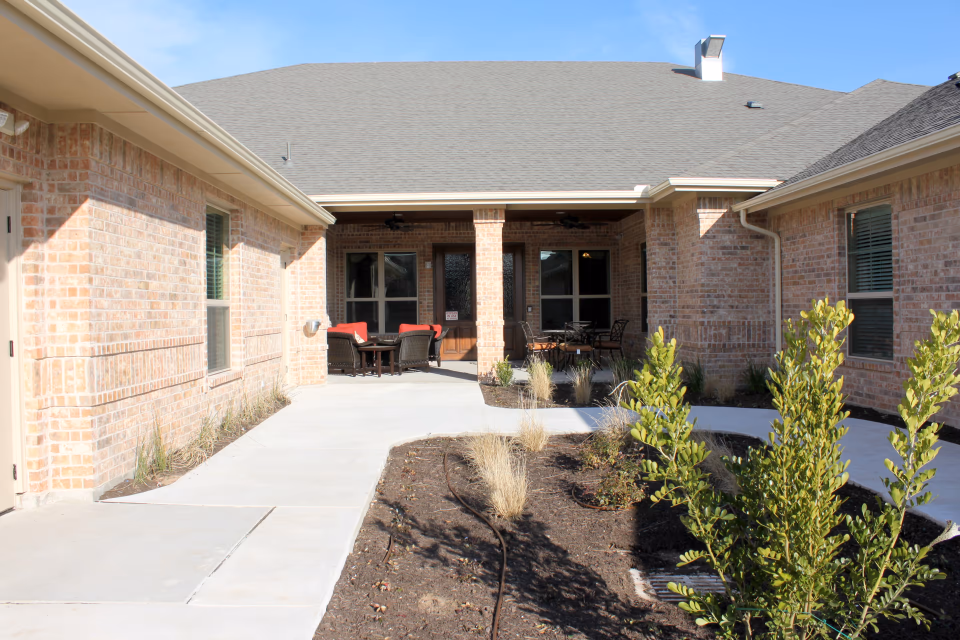 Sunlit brick courtyard with a paved walkway leading to a covered patio furnished with chairs and tables.