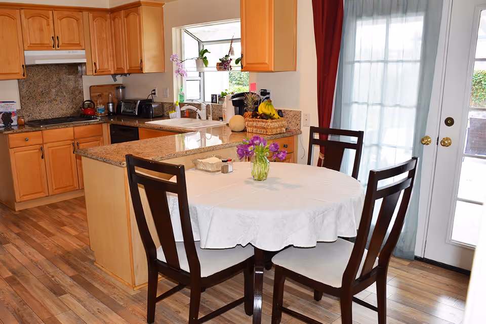 A kitchen and dining area with wooden cabinets, granite countertops, and a round dining table covered with a white tablecloth. The table has a small vase with purple flowers and is surrounded by four dark wooden chairs with light cushions. There is a basket of fruit on the counter and a glass door with sheer curtains letting in natural light.