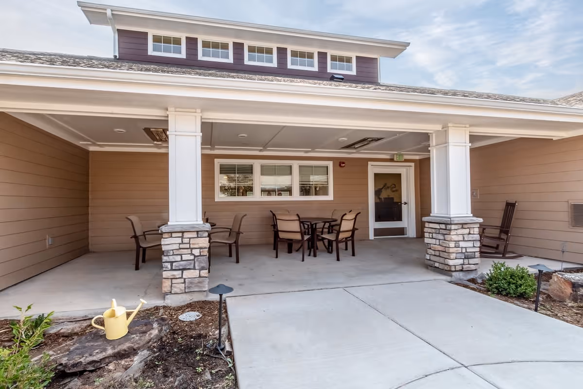 Covered outdoor patio area with a concrete floor, two stone pillars, a round table with four chairs, a rocking chair, and a yellow watering can placed on a rock in a small garden bed nearby.
