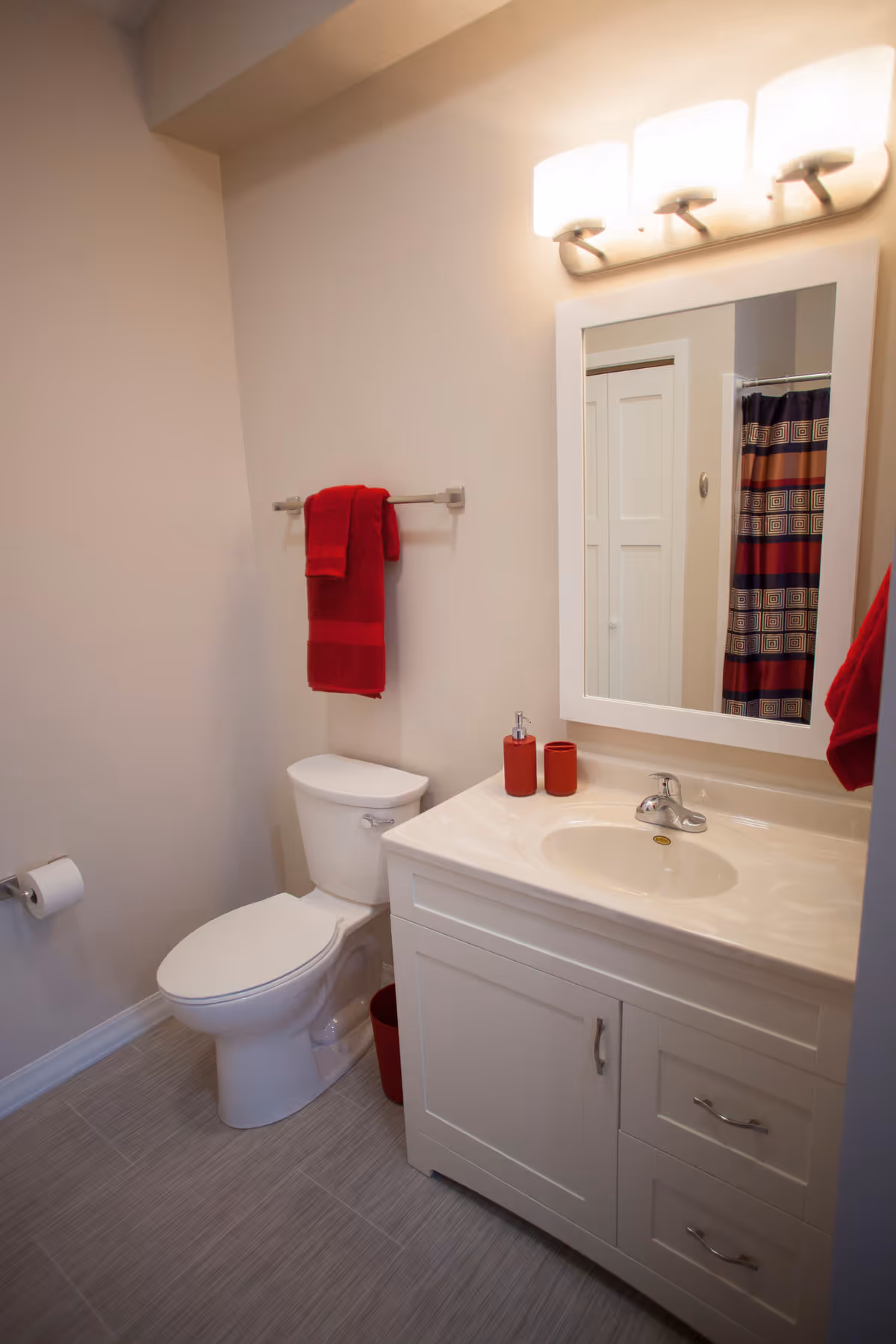 A clean bathroom with a white toilet, a white vanity with a sink, a large mirror above the sink, and a light fixture with three lights. There are red towels hanging on a towel rack and a red waste bin next to the toilet. A shower curtain with red, brown, and beige stripes is partially visible in the mirror.