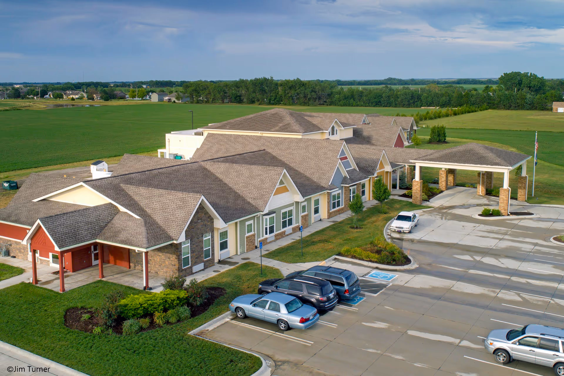 Aerial view of Sunflower Terrace, a single-story senior living facility with multiple peaked roofs and a covered entrance. The building is surrounded by green lawns and landscaping, with a parking lot in front containing several cars. The background shows open fields and a few distant houses under a partly cloudy sky.