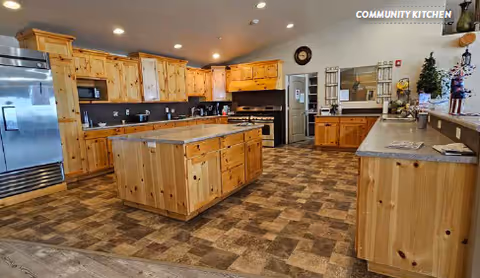 Spacious community kitchen with wooden cabinets, a central island, stainless steel refrigerator and stove, and a tiled floor.