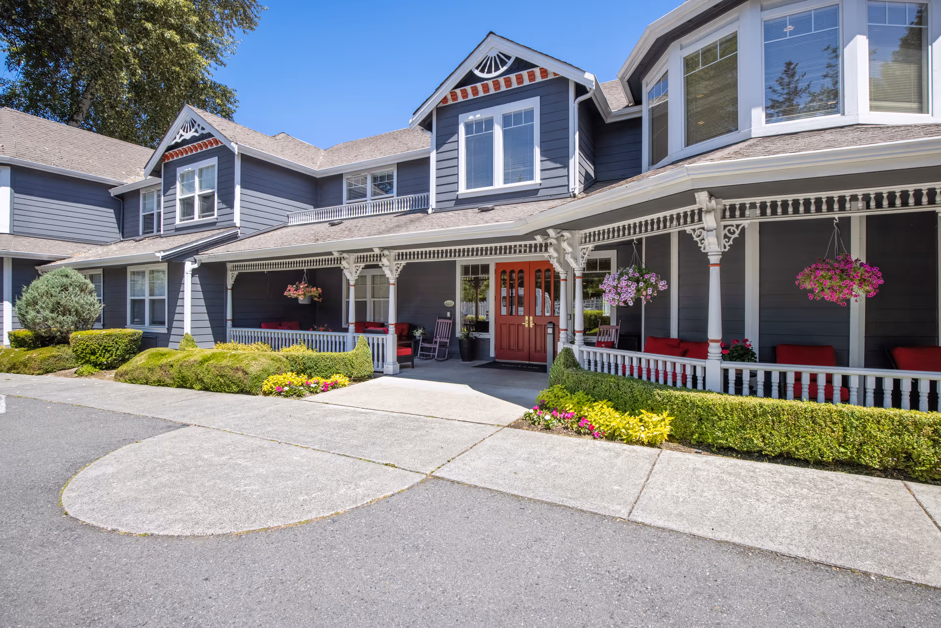 Front exterior view of a two-story building with gray siding and white trim, featuring a covered porch with red cushioned seating and hanging flower baskets. The entrance has double red doors, and there are well-maintained shrubs and flowers along the walkway under a clear blue sky.
