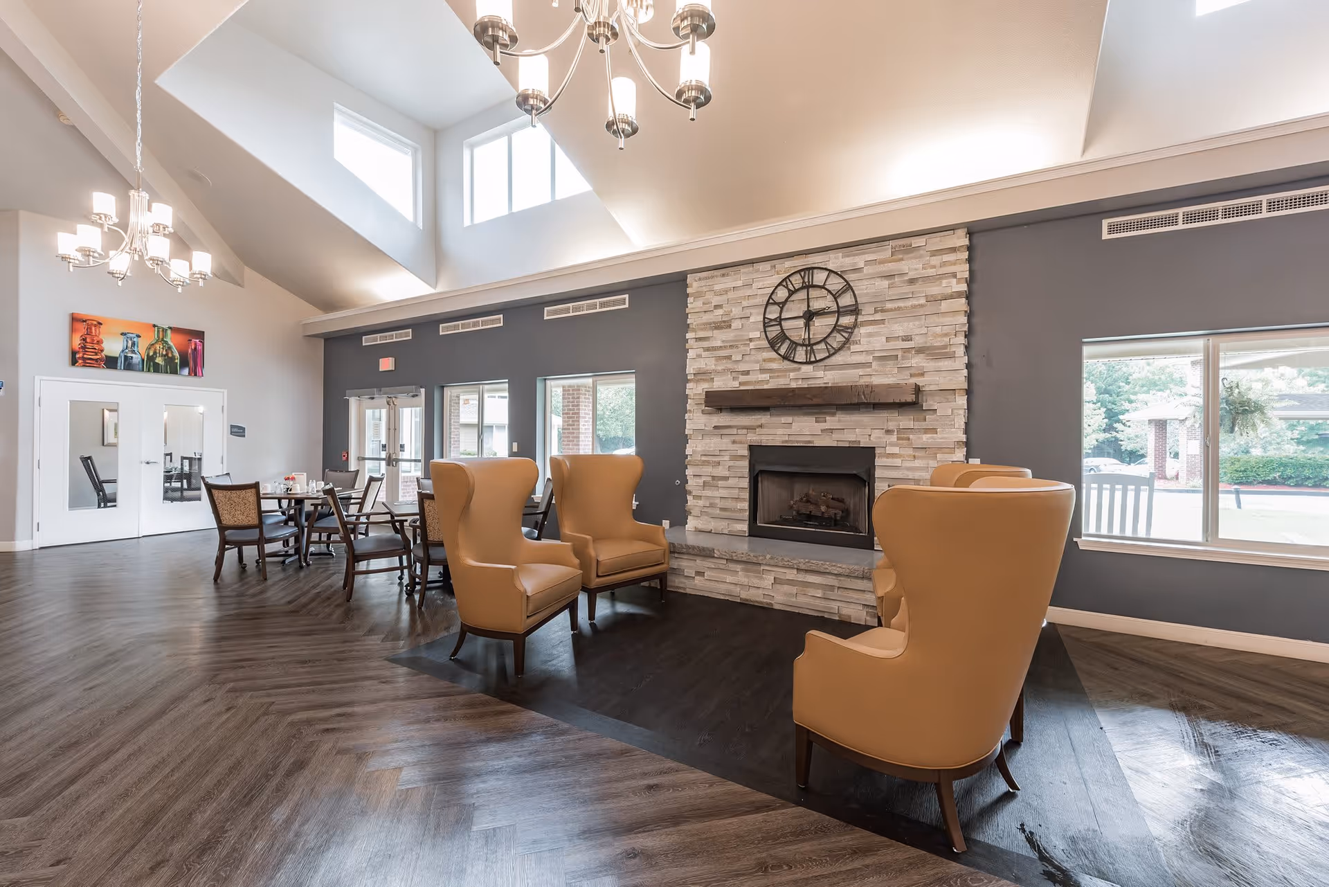 A spacious, well-lit common area with high ceilings and large windows. The room features a stone fireplace with a large clock above it, surrounded by four tan leather armchairs. In the background, there are dining tables and chairs, modern chandeliers, and double doors leading outside.