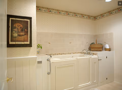 A walk-in bathtub with safety handles installed in a bathroom. The walls are decorated with light-colored wallpaper and a floral border near the ceiling. There is a framed picture on the wall and a wicker basket on a cabinet next to the tub.