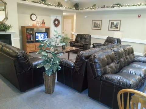 A cozy common living room with dark leather sofas and armchairs arranged around a coffee table and TV, a potted plant, framed pictures, and holiday garland along the ceiling.