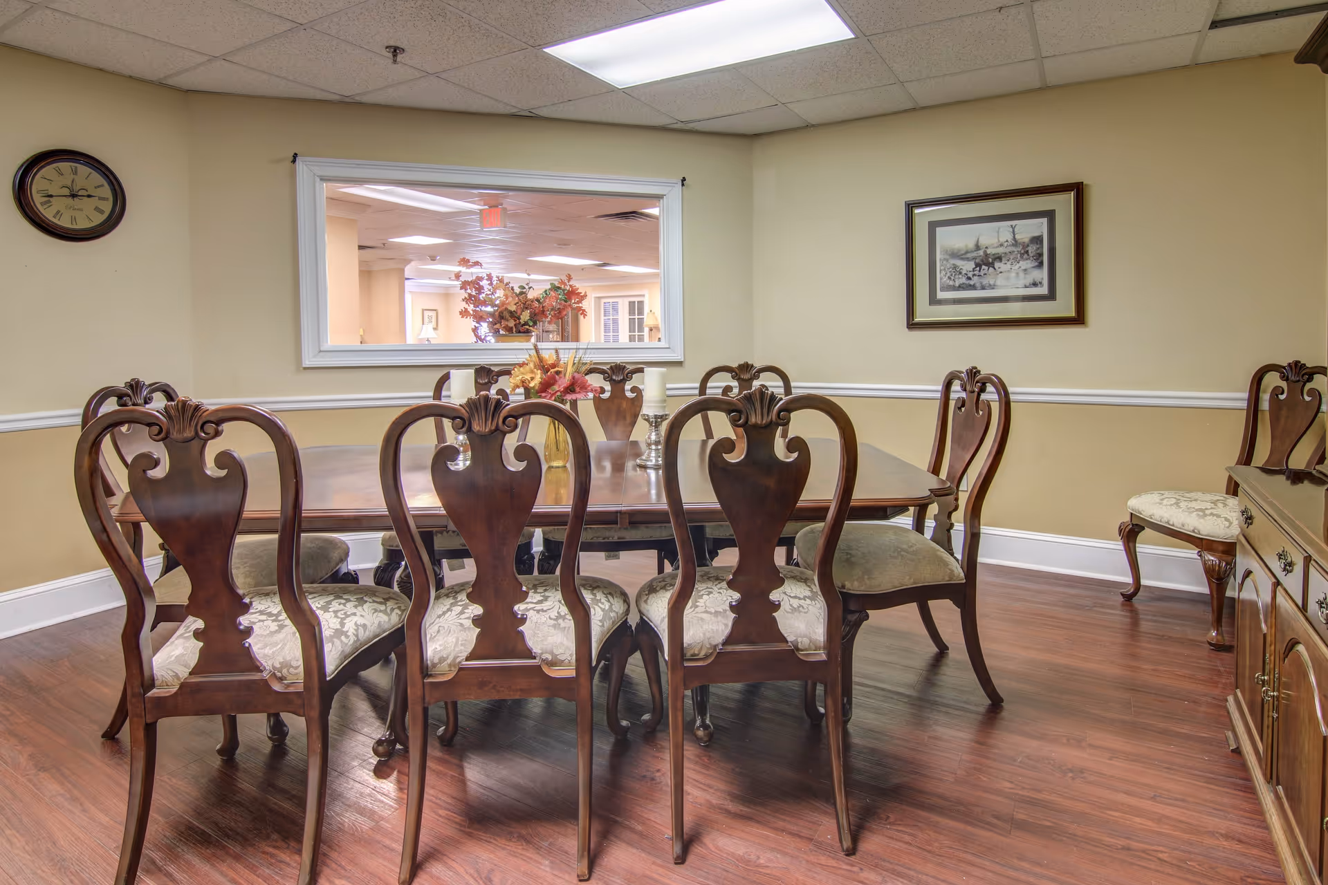 A dining room with a wooden dining table surrounded by six ornate wooden chairs with cushioned seats. The room has beige walls with white trim, a framed picture on the wall, a clock, and a large window looking into another room. There is a floral centerpiece and a candle on the table. The floor is wood.