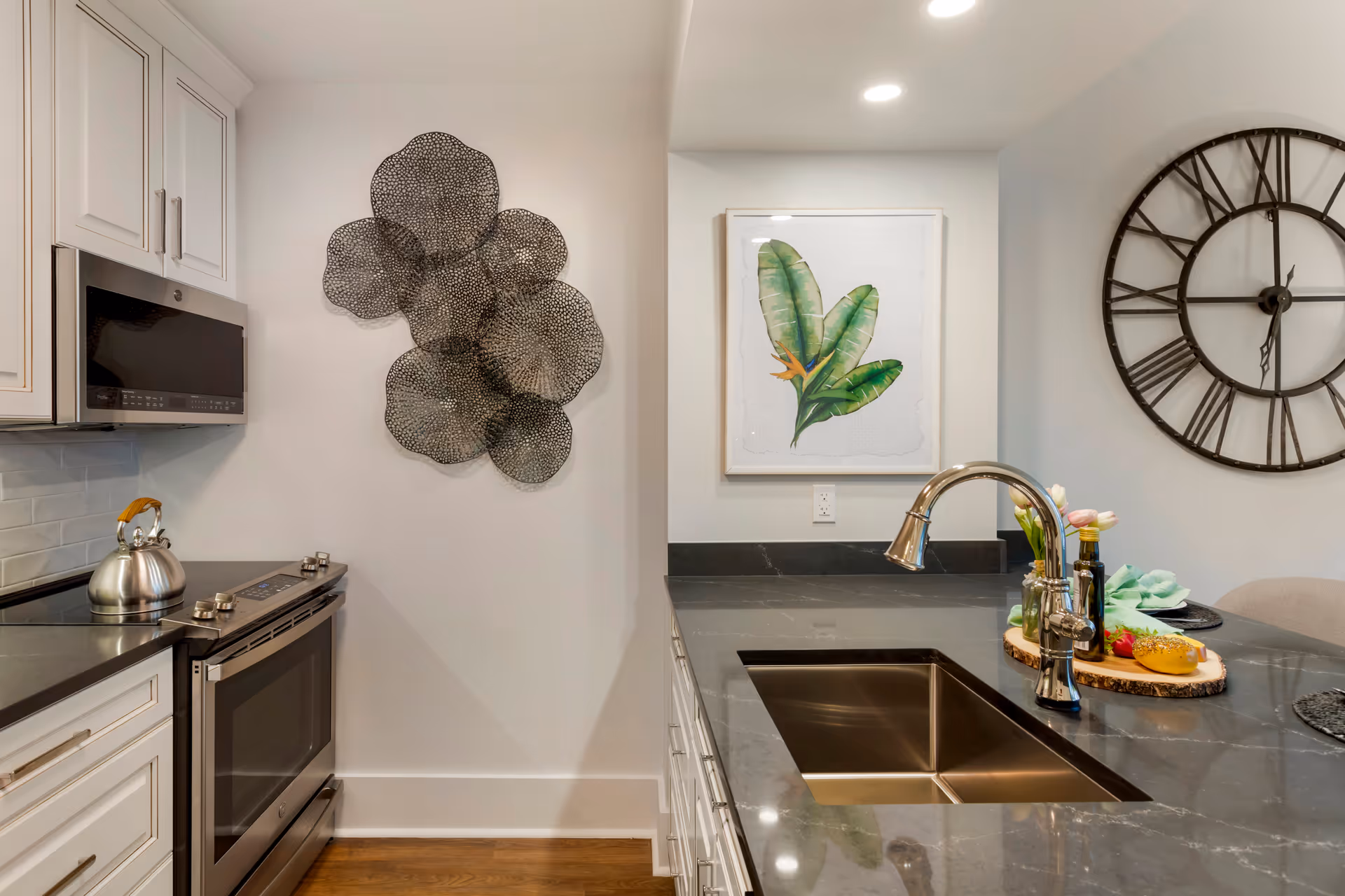Modern kitchen with white cabinetry, a stainless steel stove with a kettle, a black countertop with an integrated sink and a chrome faucet. Wall decor includes a metal abstract sculpture and a framed botanical print. A large black wall clock is visible on the right side.
