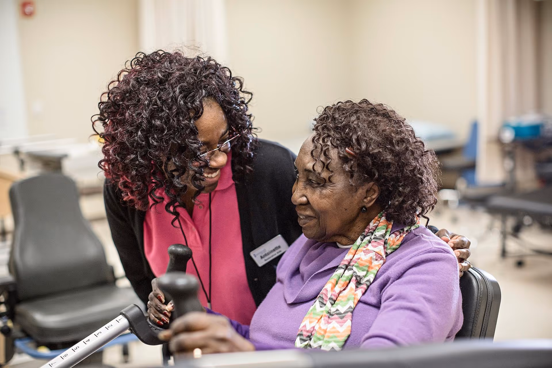A caregiver with curly hair and glasses smiles warmly at an elderly woman seated in a wheelchair. The elderly woman is wearing a purple sweater and a colorful scarf. They are in a room with medical or therapy equipment in the background.