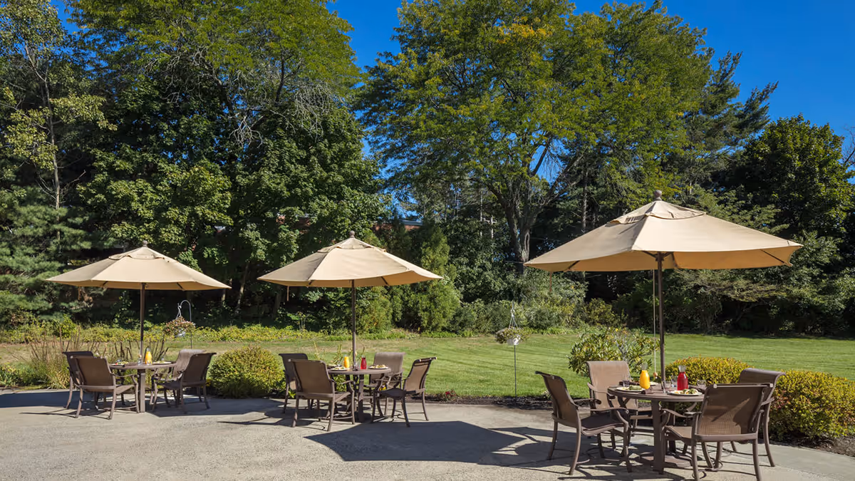 Outdoor patio area with three round tables, each shaded by a large beige umbrella. Each table is surrounded by four brown chairs. The patio overlooks a well-maintained lawn and is bordered by green bushes and tall trees under a clear blue sky.