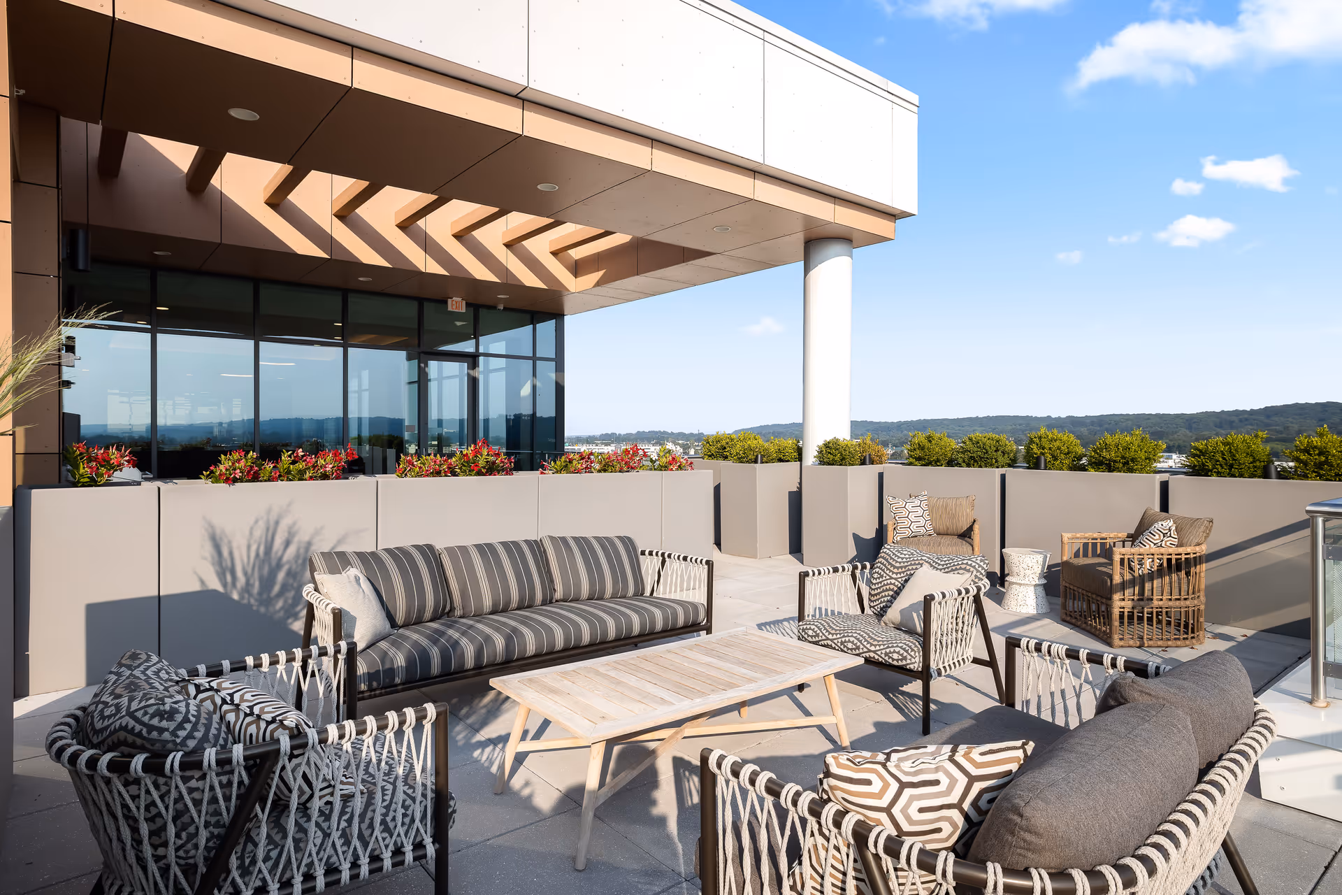 Outdoor patio area with modern wicker and cushioned seating arranged around a wooden coffee table. The patio is surrounded by planters with green shrubs and red flowers, under a partly cloudy blue sky with a view of distant hills.