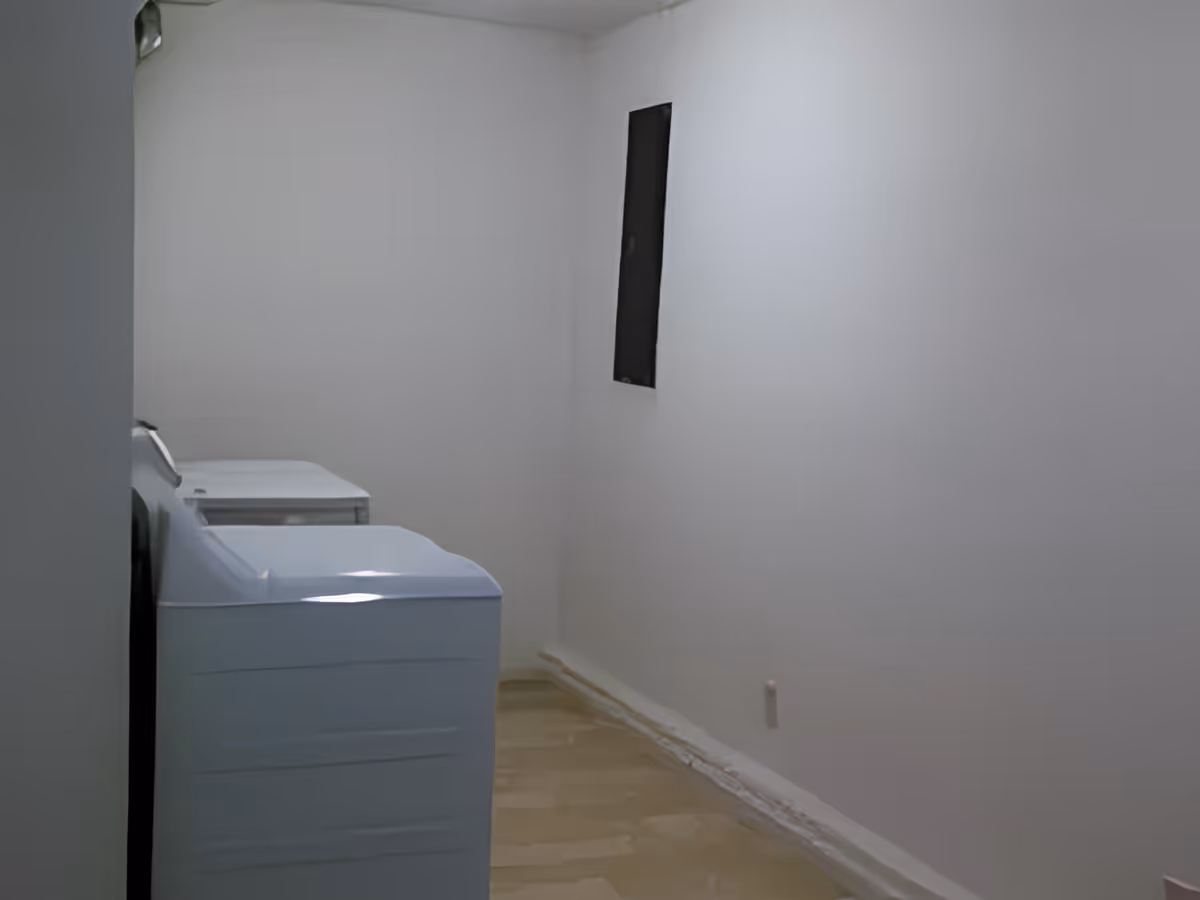 Narrow laundry/utility room with two white top-loading machines against a white wall and an unfinished floor.