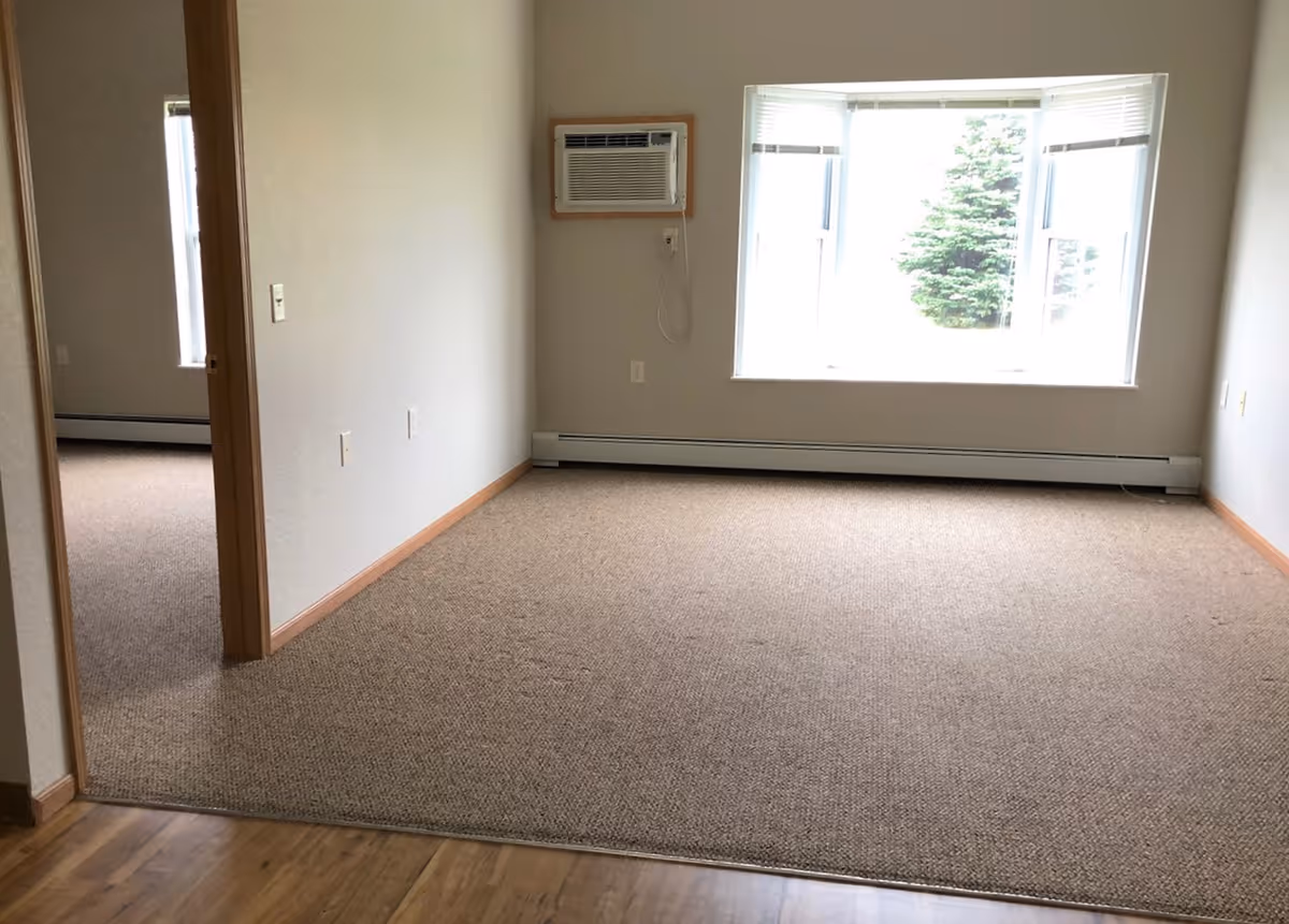Empty carpeted living room with a large window, a wall air conditioner, baseboard heating, and an open doorway to another room.