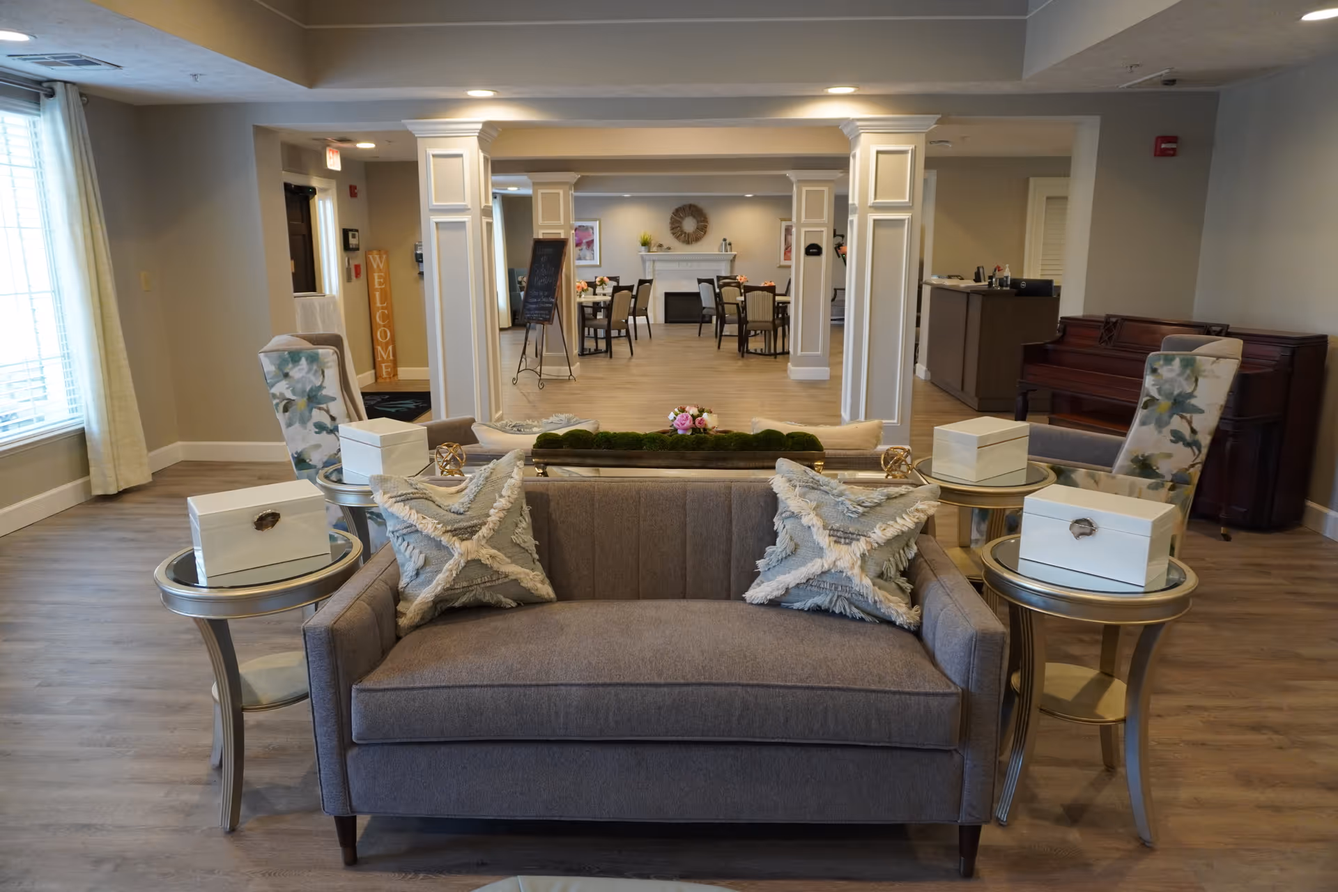 A cozy senior living facility common area with a gray sofa adorned with two decorative pillows, two round side tables each holding a white box, and two floral upholstered chairs. In the background, there is a dining area with tables and chairs, a fireplace with a wreath above it, and a piano to the right.