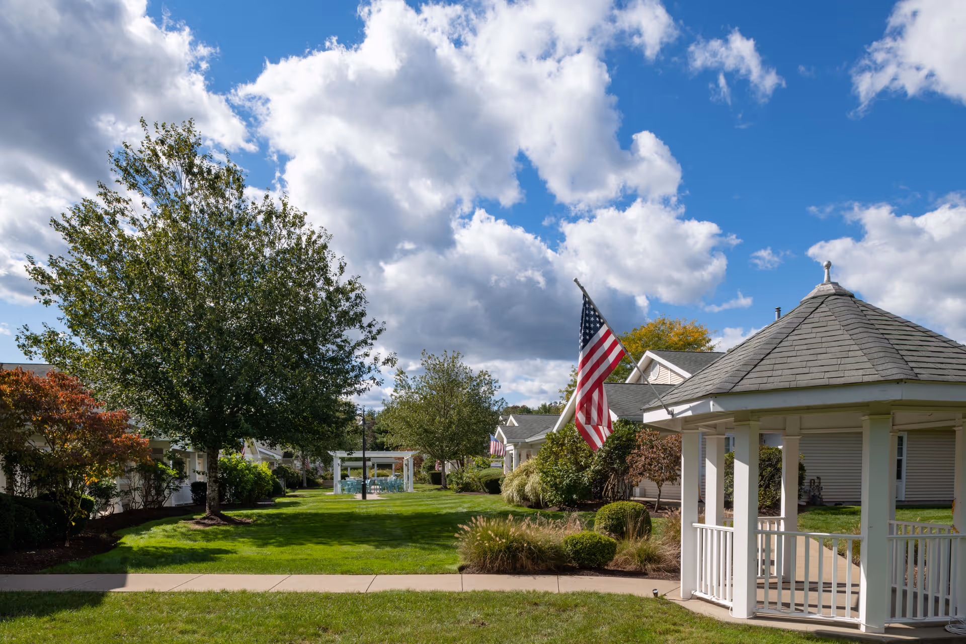 A well-maintained outdoor area at The Cottages of Dartmouth Village featuring a green lawn, trees, shrubs, a white gazebo, and American flags on poles. The sky is partly cloudy with blue patches visible.