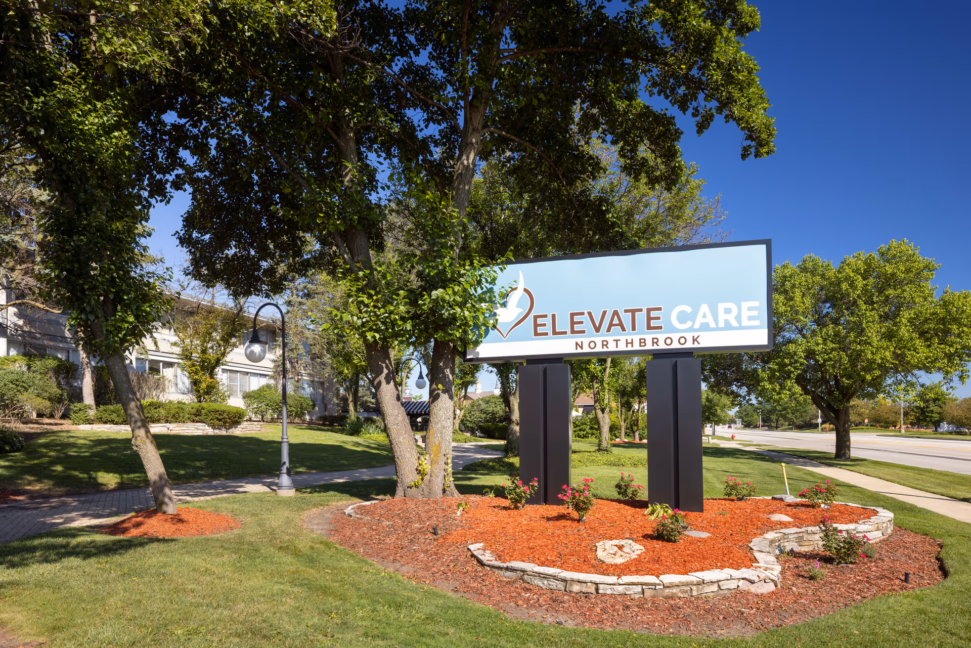 Outdoor view of the Elevate Care Northbrook facility sign surrounded by landscaped greenery, trees, and a clear blue sky.
