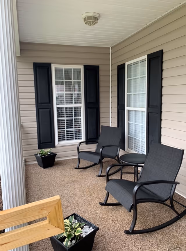 Covered outdoor porch area with two black rocking chairs and a small round black table between them. The porch has beige siding with two windows featuring black shutters. There are two black square planters with green plants, one near the windows and one near the wooden railing. A white column supports the porch roof.