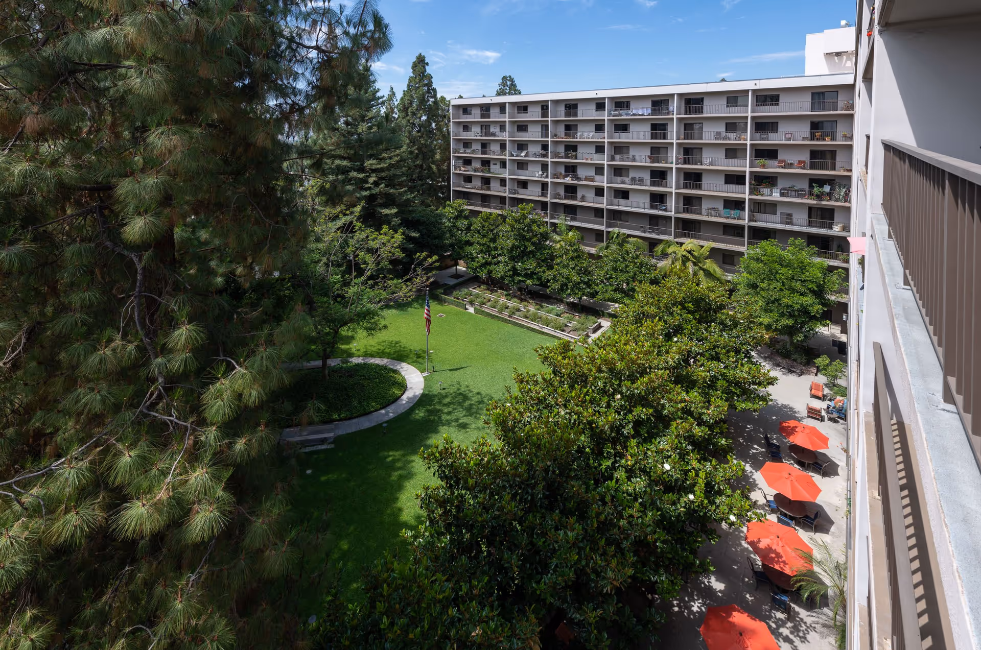 View over a green courtyard with trees, an American flag, red patio umbrellas, and a multi-story residential building with balconies.