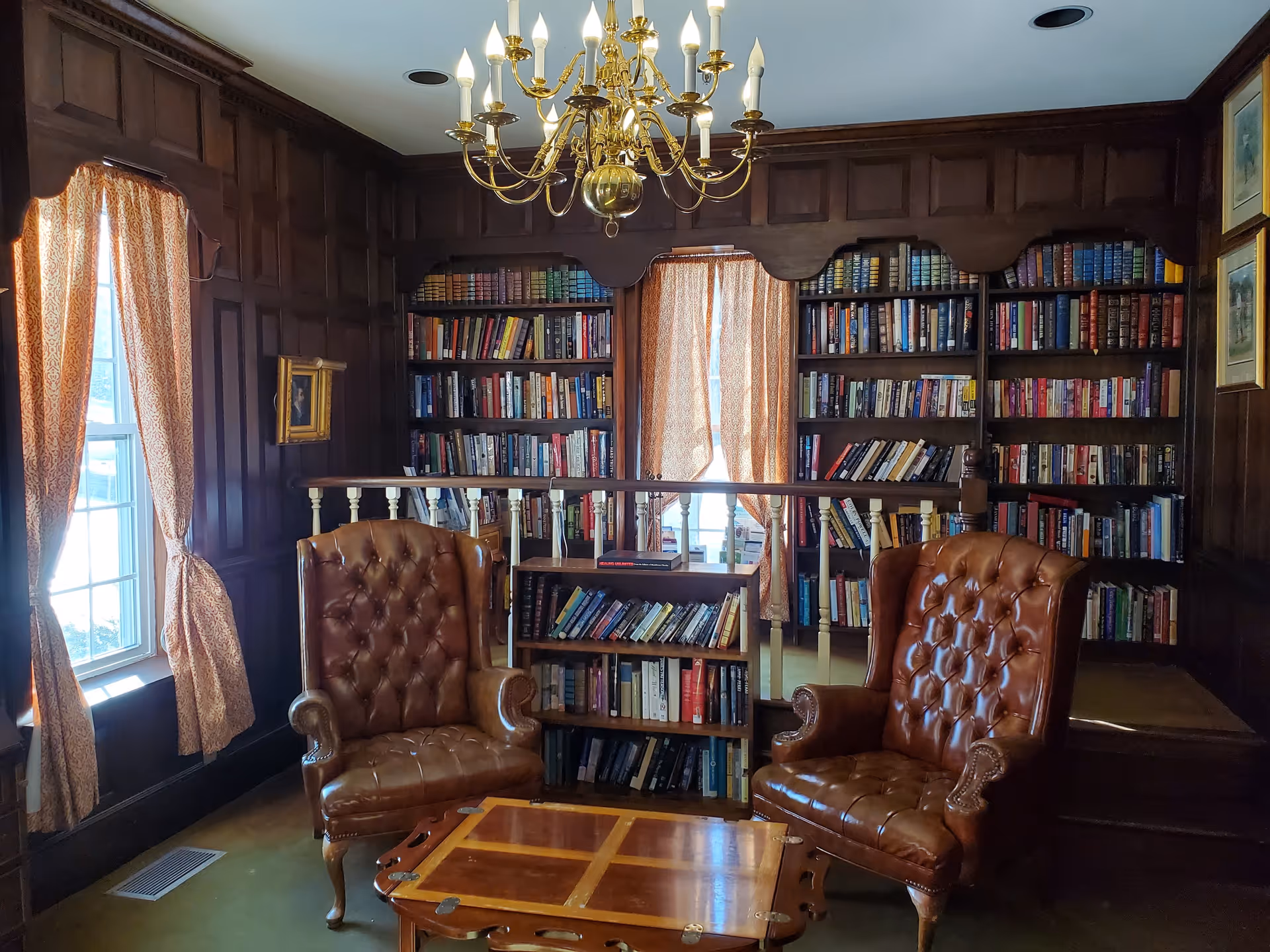 Cozy wood-paneled library room with bookshelves, two brown leather armchairs, a wooden coffee table, and a chandelier.