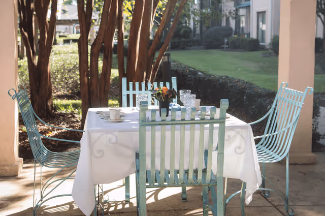 Outdoor patio area with a white tablecloth-covered table set with cups, glasses, and a small flower arrangement, surrounded by four light blue metal chairs. Trees and greenery are visible in the background.