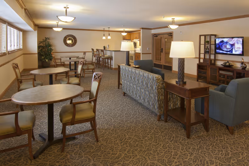 Spacious senior living common area with round dining tables, lounge seating, a TV, and a kitchenette in the background.