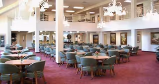 Spacious dining area with multiple round tables surrounded by green cushioned chairs, red carpet flooring, white walls, and several chandeliers hanging from the ceiling. The room has a second-floor balcony overlooking the dining space and decorative wall art.
