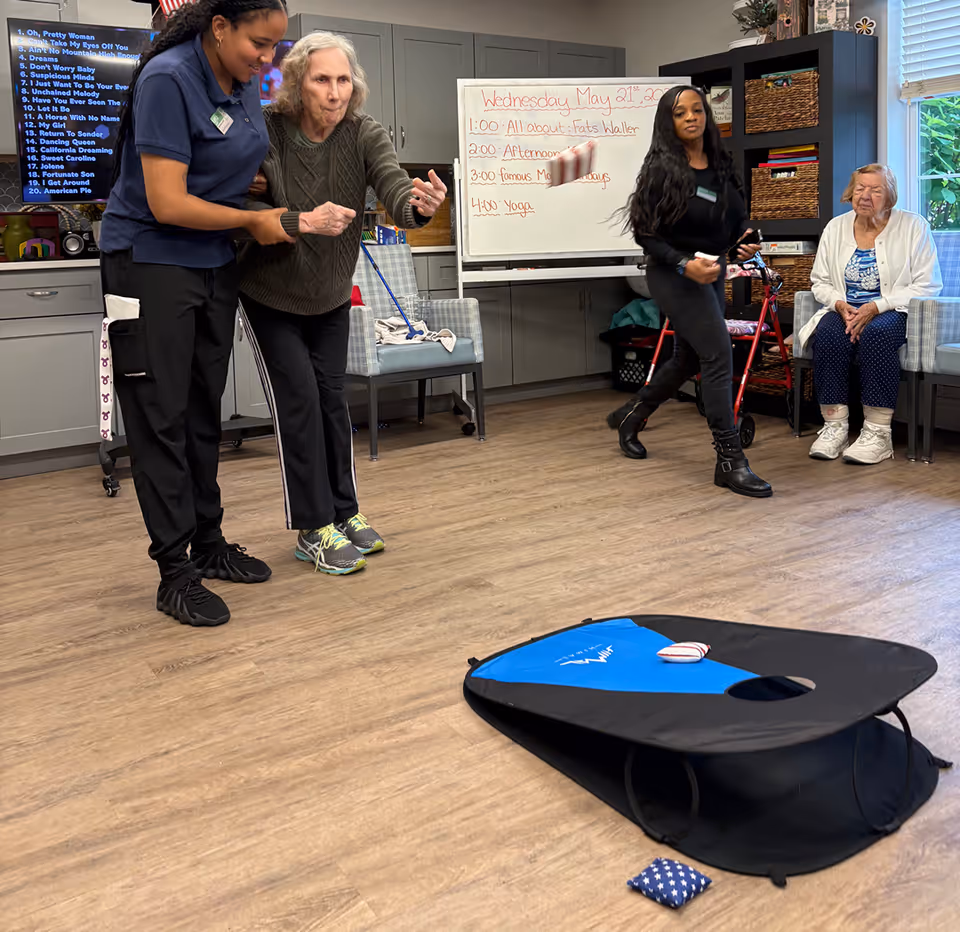 Residents and staff play a beanbag cornhole game in a senior living activity room with chairs, shelving, and a whiteboard showing the day’s schedule.