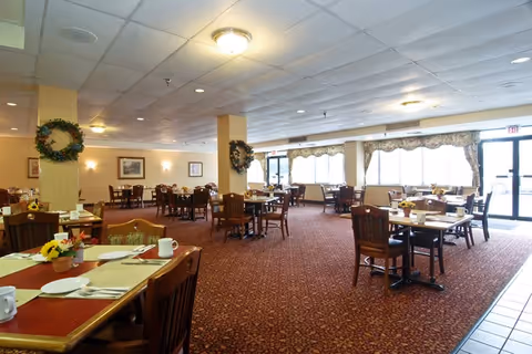 A spacious dining room with multiple wooden tables and chairs arranged neatly. Each table is set with placemats, cups, and small flower arrangements. The room has carpeted flooring, large windows with curtains allowing natural light, and decorative wreaths on the walls and columns.