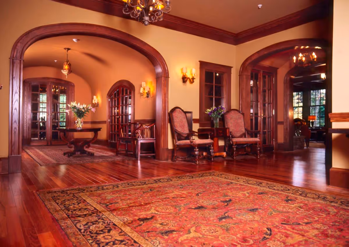 An elegant interior space with polished hardwood floors and a large ornate red and gold area rug. The room features arched doorways with dark wood trim, two upholstered chairs with a small table and flower vase between them, and a round wooden table with a flower vase in the background. Warm yellow walls are accented by wall sconces and a chandelier hanging from the ceiling.