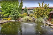 Landscaped outdoor pond with a small fountain, rocks, shrubs and trees in front of a multi-story retirement facility building.