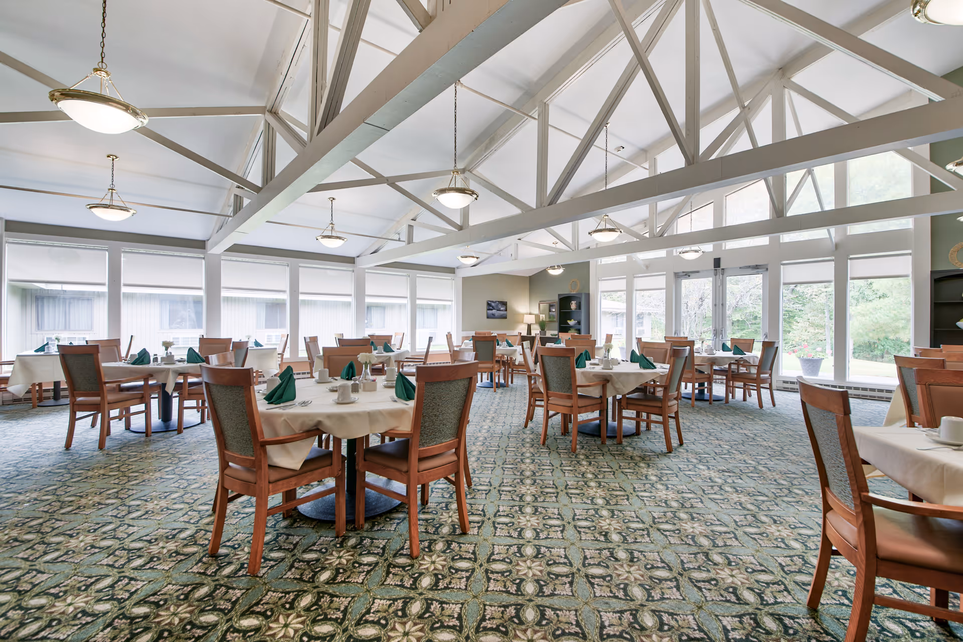 A spacious dining room with multiple round tables covered with white tablecloths and set with green folded napkins, cups, and silverware. The room features large windows allowing natural light to fill the space, a patterned carpet, and exposed white ceiling beams with hanging light fixtures.