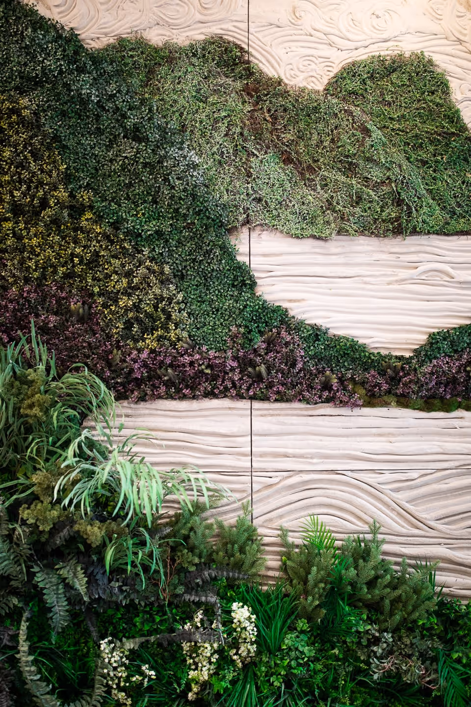 A decorative wall featuring a textured, wavy beige surface with various green and purple plants arranged artistically to create a natural, garden-like appearance.
