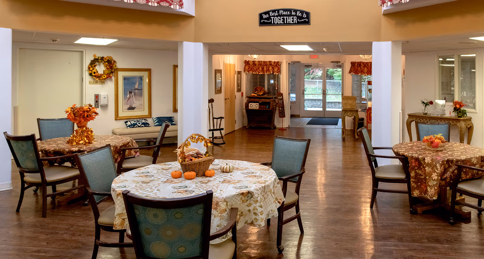 Interior view of a senior living facility common area with round tables covered in autumn-themed tablecloths and decorated with small pumpkins and fall foliage centerpieces. Several chairs surround the tables. The room has wooden flooring, a bench with cushions, framed artwork on the walls, and a sign above the entrance that reads 'The Best Place To Be Is TOGETHER'. Double glass doors lead outside, and there is a rocking chair and a small table with flowers near the entrance.