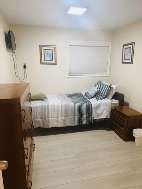 Small furnished single bedroom with a twin bed dressed in striped bedding, a dresser, nightstand, wall-mounted TV and framed pictures.