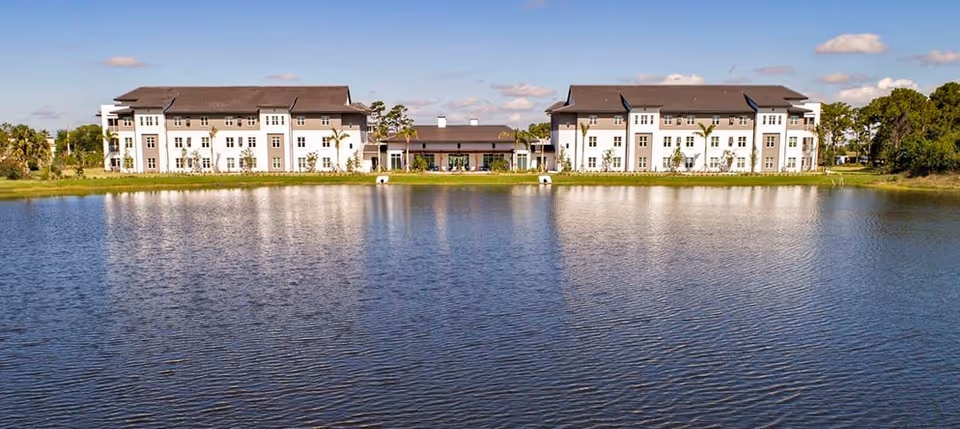 Three-story senior living building complex reflected across a large lake under a blue sky.